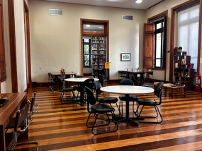 Vista da sala de leitura do Centro Cultural Justiça Federal, com ambiente amplo e iluminado. O espaço tem piso de madeira e paredes claras com detalhes em madeira. Mesas redondas e cadeiras estão organizadas pelo salão, algumas com pessoas sentadas estudando ou lendo. Ao fundo, há estantes e portas altas de madeira, além de janelas que permitem a entrada de luz natural, criando um clima tranquilo e acolhedor.