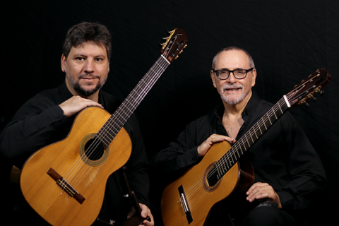 A imagem mostra dois homens sentados, cada um segurando um violão clássico. Ambos vestem camisas pretas e estão de frente para a câmera, com expressões serenas. O homem à esquerda tem barba curta e cabelo castanho, enquanto o da direita usa óculos, tem barba grisalha e está sorrindo. O fundo é preto, destacando os músicos e seus violões. A imagem transmite um clima de seriedade e paixão pela música.