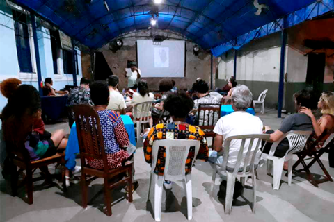 A imagem mostra várias pessoas sentadas em cadeiras de diferentes tipos, olhando para uma tela de projeção em um ambiente coberto, com um teto azul. A sala parece ser um espaço simples e comunitário, com paredes parcialmente desgastadas. Na tela, há uma apresentação, mas o conteúdo não está claro. A iluminação é suave, com foco na tela, e o ambiente sugere uma reunião ou evento informativo.