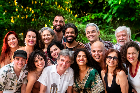 Na foto, um grupo de 15 pessoas, entre homens e mulheres das mais variadas faixas etárias, sorri para a câmera. Eles estão em uma área aberta, com vegetação ao fundo. Há mulheres de cabelos castanhos lisos e enrolados, ruiva, e grisalhas; homens de cabelos castanhos curtos, castanhos enrolados, careca e grisalhos. As roupas se misturam entre estampadas e lisas (brancas e pretas). É possível perceber a ponta de um violão na mão de um dos homens que está posicionado ao centro, na fileira da frente da foto.