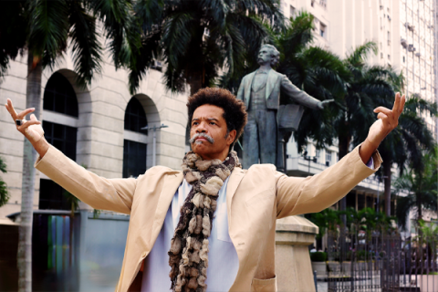 A foto mostra um homem negro de bigode grisalho e cabelos afro com os braços erguidos. Usa blazer bege claro, blusa branca e cachecol em tons marrons. Ele está com um olhar obstinado, à frente de uma estátua de outro homem de bigode e paletó. Há palmeiras (árvores) e prédios no cenário.