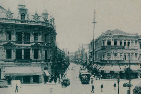 Fotografia antiga do Largo da Carioca, Rio de Janeiro. Os edifícios em destaque possuem arquitetura elaborada com ornamentos detalhados nas fachadas, característica do estilo europeu do início do século XX. A rua é movimentada, com pessoas caminhando e alguns veículos de tração animal. Há também uma linha de bonde no centro da via. O cenário transmite uma atmosfera clássica e urbana, com foco na vida cotidiana da época.