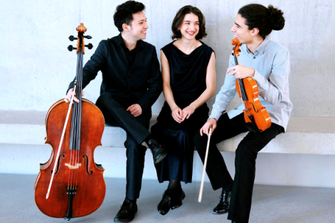 Na foto, dois homens e uma mulher branca estão sentados em um banco; todos sorriem. Um deles (à esquerda) usa paletó preto e segura um violoncelo. A mulher, no meio dos dois, veste vestido longo preto e olha para um dos musicistas. O homem da direita usa blusa social clara e calça preta; ele segura um violino.