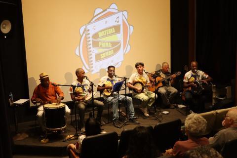 Um grupo de músicos, se apresentam em um palco. Todos estão sentados, tocando e cantando. Entre os instrumentos musicais, estão: pandeiro, banjo, violão, cavaquinho e tan tan.