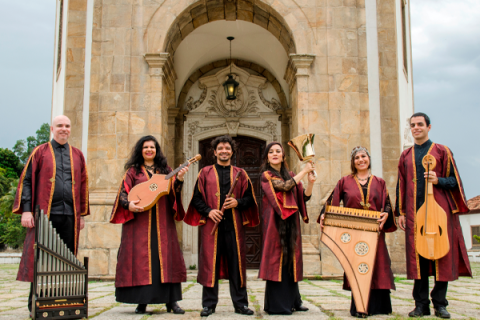 A imagem mostra um grupo de seis músicos, todos vestidos com trajes tradicionais, longos e em tons de vinho com detalhes dourados. Eles estão posando em frente a um edifício antigo, que parece ser uma igreja com uma arquitetura clássica de pedra. Cada um está segurando um instrumento musical antigo: alaúdes, flauta, um órgão portátil, uma harpa e um par de sinos. O grupo aparenta ser especializado em música antiga ou medieval, e o cenário reforça essa atmosfera histórica e cultural.