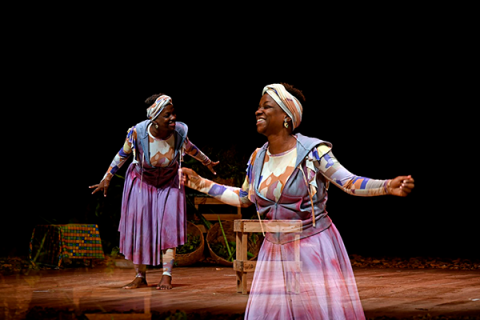 A imagem mostra um palco com uma mulher negra sorridente, descalça, usando vestido longos roxos e faixa de cabeça combinando – parece estar em uma performance teatral, com gestos abertos e expressões alegres. Esta mesma imagem da mulher é espelhada e sobreposta em um primeiro plano da fotografia, com certa transparência. O cenário tem um fundo escuro e alguns objetos de madeira, sugerindo uma ambientação simples e natural. Há uma sensação de movimento e energia, destacando a leveza e o dinamismo da cena.