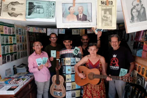 Grupo com 5 homens e uma mulher brancos, em sala com livretos de literatura de cordel. Um deles veste traje plástico transparente com livretos amarelos, azuis e brancos, de cordel. Usa chapéu preto e tem um violão em uma das mãos. Uma mulher de óculos, ao centro, toca outro violão e veste vestido estampado na cor vinho. Os outros homens seguram livretos de cordel, alguns usam óculos e alguns possuem cabelos grisalhos curtos. No 1º plano, na parte superior da imagem, há 5 livros do tema pendurados em varal.