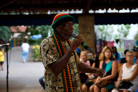 A imagem mostra um homem negro segurando um microfone enquanto fala ou se apresenta para uma plateia. Ele está usando uma boina de crochê nas cores verde, amarelo e vermelho, além de um cachecol listrado nas mesmas cores. Veste uma camisa estampada com desenhos de vasos. Ao fundo, há várias pessoas sentadas, ouvindo atentamente. O ambiente parece ser ao ar livre, com vegetação ao fundo, e o clima sugere um evento cultural ou social.