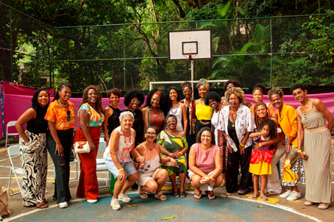 A imagem mostra um grupo de aproximadamente 25 mulheres posando juntas em uma quadra de esportes ao ar livre cercada por uma grade e vegetação ao fundo. Elas estão sorrindo e parecem celebrar ou participar de um evento social. Algumas usam roupas coloridas e estampadas, enquanto outras vestem roupas mais neutras. Entre elas, há uma criança e mulheres de diferentes idades. Ao fundo, há uma tabela de basquete. O ambiente é acolhedor e de camaradagem, com um clima de união.