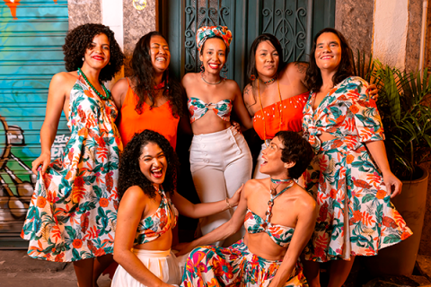 A imagem mostra um grupo de sete mulheres sorrindo e posando juntas em frente a uma porta, em ambiente externo. Elas usam roupas coloridas com estampas tropicais e vibrantes, predominando tons de laranja, verde e branco. Alguns vestem vestidos, enquanto outros usam tops e saias, criando uma estética alegre e descontraída. O cenário urbano e a expressão das mulheres passam uma sensação de celebração e camaradagem.