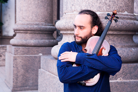 A imagem mostra um homem com cabelo preso e barba, usando uma camisa azul, abraçando um violino. Ele olha para o lado, com uma expressão contemplativa. Ao fundo, há uma estrutura de pedra, sugerindo que ele está em um local histórico ou cultural. A cena transmite uma sensação de apreço pela música e pelo instrumento, em um ambiente tranquilo e introspectivo.