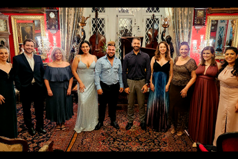 Grupo de pessoas posando em um ambiente elegante e decorado de forma clássica, com paredes vermelhas, lustres, quadros e mobília detalhada. Todos estão vestidos formalmente, com trajes de gala como vestidos longos e ternos, sugerindo que estão participando de um evento especial ou cerimônia. O ambiente tem um tapete com padrão floral e móveis sofisticados, criando uma atmosfera luxuosa e histórica. Ao fundo, há detalhes arquitetônicos, como janelas em estilo gótico e estatuetas decorativas