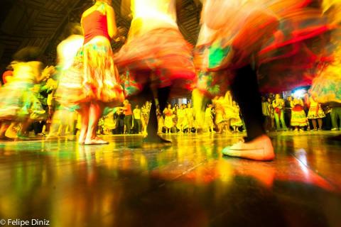 A imagem mostra uma cena dinâmica de dança em grupo, com pessoas girando em um salão iluminado por luzes amarelas e coloridas, criando um efeito de movimento borrado. Os dançarinos vestem roupas tradicionais, como saias rodadas e coloridas, típicas de danças folclóricas brasileiras. O reflexo das cores no piso contribui para uma atmosfera vibrante e festiva. A foto, assinada por Felipe Diniz, capta a energia e a vivacidade da dança popular.