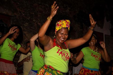 A imagem mostra uma mulher sorridente no centro, usando uma camiseta verde com o logo "Zanzar" em vermelho, acompanhada por outras mulheres ao fundo, também vestindo camisetas iguais. Ela usa uma saia estampada e um lenço colorido na cabeça, enquanto dança com os braços erguidos em um ambiente descontraído. O fundo parece ser um espaço interno com iluminação baixa e detalhes de pedra ou tijolos. Outras pessoas ao redor também estão interagindo e se divertindo. A cena transmite alegria e celebração.