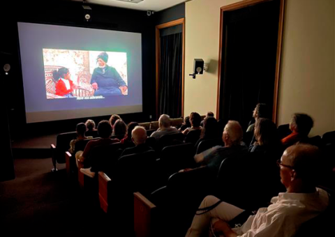 A imagem mostra um grupo de pessoas assistindo a um filme em uma pequena sala de cinema ou auditório. A projeção na tela exibe uma cena de diálogo entre uma menina e um senhor de turbante, com legendas visíveis na parte inferior. A sala está escura, com as luzes apagadas, criando um ambiente imersivo para os espectadores, que estão sentados em fileiras de cadeiras acolchoadas. Todos estão voltados para a tela, e o clima parece tranquilo e focado na exibição do filme.