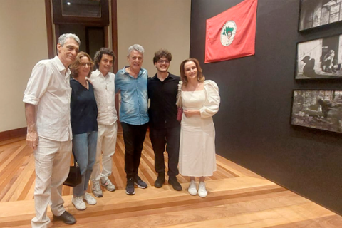 A imagem mostra um grupo de seis pessoas (entre elas o cantor Chico Buarque, a diretora-geral do CCJF, o curador e os artistas das exposições) posando na exposição Lona Preta: o MST no Olhar de Francisco Proner. O espaço tem paredes escuras e piso de madeira clara. Eles estão sorrindo e usam roupas casuais, predominantemente em tons claros. Ao fundo, há uma parede com fotografias em preto e branco e uma bandeira vermelha com um símbolo verde e branco. A expressão das pessoas sugere satisfação e camaradagem.