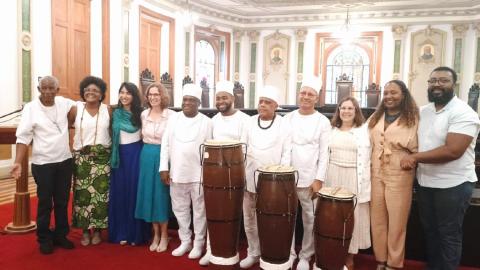 A imagem mostra um grupo de pessoas posando juntas em um ambiente elegante, com arquitetura clássica, incluindo colunas e detalhes decorativos nas paredes. Algumas pessoas vestem roupas brancas tradicionais e estão ao lado de tambores, sugerindo uma conexão com práticas culturais ou religiosas. Outras estão em trajes formais ou coloridos, sorrindo. Ao fundo, há janelas com vitrais e uma atmosfera de celebração ou cerimônia.