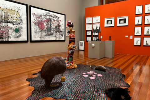 Interior de galeria de arte. No centro, há uma instalação artística composta por tapete com padrão geométrico, esculturas de animais, como um pássaro marrom, um tatu e a metade inferior de uma capivara com diversas facas cravadas no local de onde seria a cabeça, além de outros objetos inusitados. Ao fundo, quadros grandes com arte abstrata, pendurados na parede cinza. À direita, uma parede laranja exibe quadros menores em arranjo ordenado. O piso de madeira polida dá ao espaço um ar sofisticado e acolhedor.