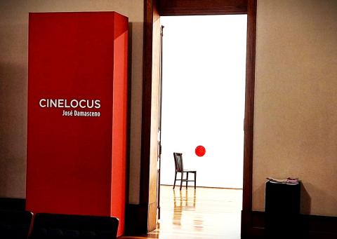 A imagem mostra a entrada de uma exposição chamada "Cinelocus" do artista José Damasceno. À esquerda, há um grande painel vermelho com o nome da exposição e do artista em letras brancas. Ao centro, uma porta aberta revela um espaço iluminado e minimalista, onde se vê uma cadeira preta e um pequeno círculo vermelho na parede branca. O chão é de madeira brilhante, refletindo os objetos. O ambiente sugere um cenário artístico e conceitual.
