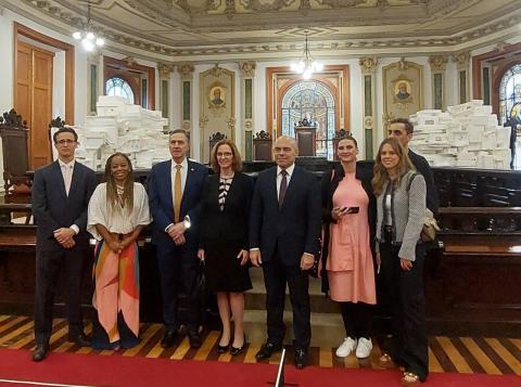 A imagem mostra um grupo de oito pessoas posando para uma foto em um salão com arquitetura clássica, repleto de detalhes ornamentais. O fundo possui um grande número de caixas brancas empilhadas e vitrais decorativos. As pessoas estão vestidas com trajes formais e semiformais. Entre elas, há homens de terno e gravata, e mulheres com vestidos e roupas casuais elegantes. O ambiente tem um ar solene, sugerindo uma visita institucional ou evento oficial.