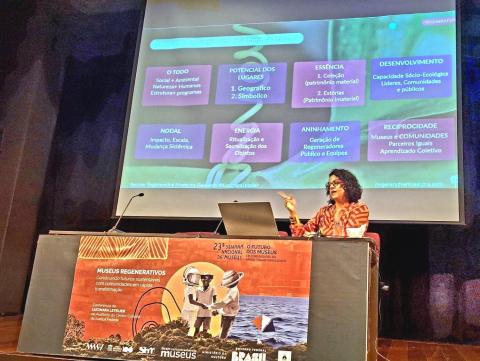 Imagem de uma palestra sobre “Museus Regenerativos” em um auditório. Uma mulher com cabelo cacheado, usando blusa vermelha estampada, está sentada à mesa com um microfone, notebook e xícara. Atrás dela, um slide apresenta conceitos como “O Todo”, “Essência” e “Reciprocidade”. Na frente da mesa, há um cartaz do evento “23ª Semana Nacional de Museus” com a frase “Construindo futuros sustentáveis com comunidades em rápida transformação”.