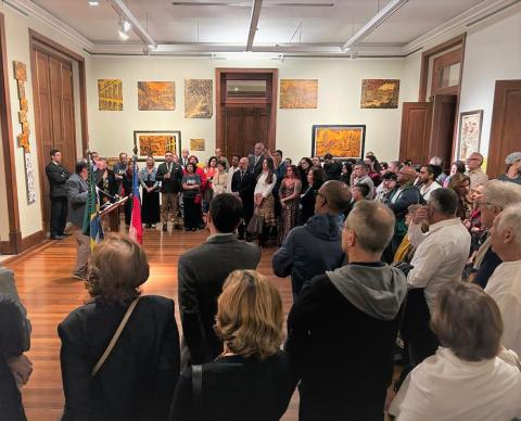 A imagem mostra uma sala de exposições cheia de pessoas, durante uma cerimônia de abertura. À esquerda, um homem fala ao microfone ao lado das bandeiras do Brasil e do Chile. O público, em pé, observa com atenção. Nas paredes, há quadros com paisagens urbanas e cores quentes. O ambiente tem piso de madeira e iluminação direcionada, transmitindo um clima solene e artístico.