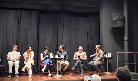 A imagem mostra um painel com seis pessoas sentadas em cadeiras no palco de um teatro, com cortinas pretas ao fundo. Três homens e uma mulher ao centro conversam animadamente, um deles segurando um microfone. Nas extremidades, dois homens observam. Há copos sobre mesas pequenas e uma pessoa do público tira uma foto na frente do palco.