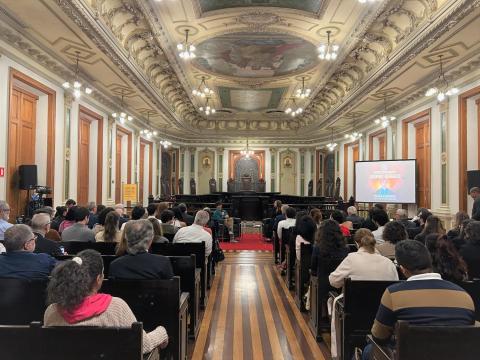 Auditório nobre com teto ornamentado e afrescos, paredes com molduras e luminárias pendentes. O espaço está cheio de pessoas sentadas, voltadas para o palco ao fundo, onde ocorre uma palestra. À direita, há um telão com a imagem do jornalista Guga Chacra e os dizeres “Encontros Acadêmicos”. O ambiente é solene e bem iluminado.