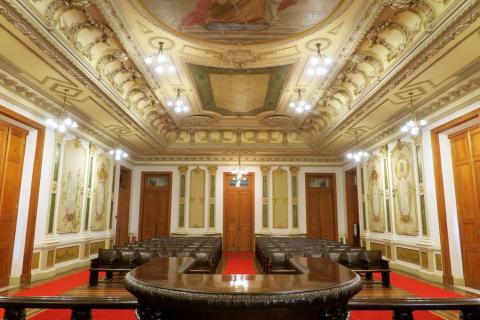 Sala de sessões com decoração luxuosa em estilo clássico. O teto é ornamentado com molduras elaboradas, pinturas e lustres pendentes. As paredes laterais exibem painéis decorativos com figuras femininas emolduradas. No centro, há uma mesa em formato de U feita de madeira escura com cadeiras estofadas ao redor. Ao fundo, poltronas organizadas em fileiras para o público. As portas e detalhes em madeira completam o ambiente.
