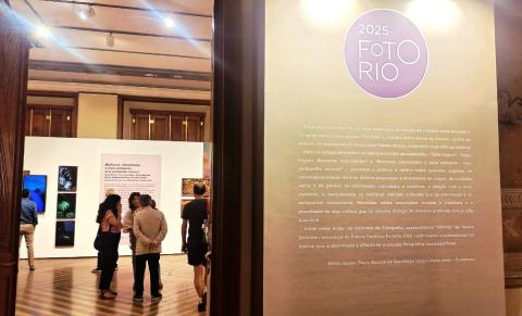 A imagem mostra uma exposição de fotografia chamada "FotoRio 2025", realizada em um espaço interno com piso de madeira e iluminação de teto. Visitantes observam painéis com fotos coloridas e textos explicativos. Na parede à direita, há um painel com o logotipo do evento e um texto introdutório sobre as temáticas abordadas na exposição, como cultura, identidade, gênero e meio ambiente.