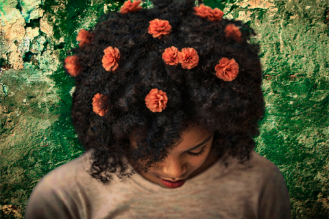 Na imagem, há uma mulher com cabelos cacheados volumosos e flores pequenas, laranjas, distribuídas pelo cabelo. Ela está inclinando a cabeça para baixo, e seu rosto está parcialmente coberto pelo cabelo. O fundo da imagem é uma parede desgastada, com textura, de tons verdes e amarelados, criando um contraste marcante com o cabelo escuro e as flores coloridas.