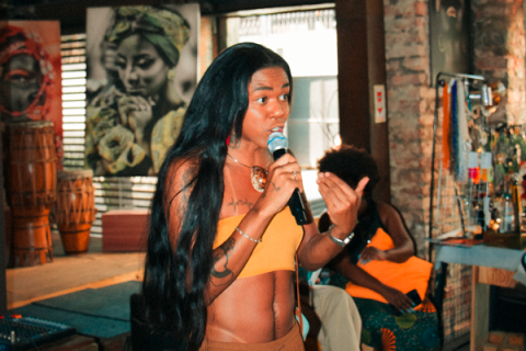 A imagem mostra uma mulher negra de cabelo comprido, liso e escuro, usando um top amarelo e segurando um microfone enquanto fala. Ela tem uma tatuagem visível no braço esquerdo e acessórios nos braços e no pescoço. Ao fundo, há uma decoração rústica com tambores e quadros, incluindo um grande retrato de uma mulher com um turbante. A atmosfera parece ser a de um evento cultural ou artístico em um espaço acolhedor, com outra mulher negra sentada no fundo.