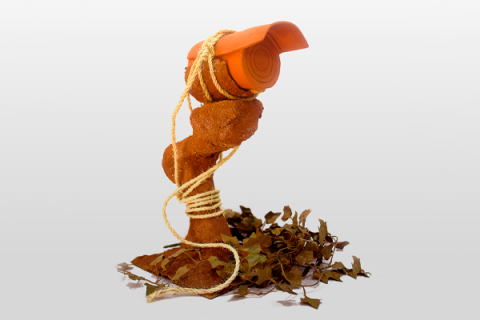 A imagem mostra um objeto semelhante a uma câmera de vigilância que parece ser feita de E.V.A laranja enrolado e amarrado, por uma corda, a um tronco ou um objeto rústico de barro. Há folhas secas espalhadas na base, criando um ambiente natural. O objeto parece representar uma fusão de tecnologia (câmera) com elementos da natureza, sugerindo um contraste entre o artificial e o orgânico.