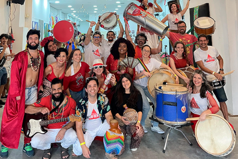 A imagem mostra um grupo alegre de pessoas em um espaço interno, segurando instrumentos de percussão como surdos, tamborins e chocalhos. Algumas vestem fantasias e roupas coloridas, criando um clima festivo. Todos sorriem e posam para a foto, transmitindo um ambiente de celebração, possivelmente de uma escola de samba ou grupo de música brasileira. Ao fundo, a decoração é simples e colorida, reforçando a atmosfera vibrante