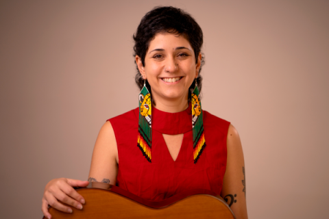 A imagem retrata uma mulher sorridente em um fundo neutro. Ela usa um vestido vermelho e ostenta longos brincos artesanais com motivos indígenas, destacando cores vibrantes como amarelo, verde e preto, e figuras de onças. Seu cabelo é curto e ondulado, e ela possui uma tatuagem visível no braço esquerdo e um piercing no nariz. A mulher está segurando um violão, sugerindo uma conexão com a música ou a arte. A expressão dela transmite simpatia e confiança.