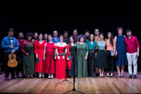 A imagem apresenta um grupo de artistas posando juntos em um palco. Todos estão bem arrumados, com roupas elegantes e coloridas, predominando tons de vermelho, verde e preto. O grupo é diverso, composto por homens e mulheres, e transmite um clima de celebração artística ou conclusão de uma apresentação. Ao fundo, um fundo preto destaca os integrantes, enquanto à frente, é possível notar um microfone no pedestal, sugerindo que o evento está relacionado à música ou performance vocal.