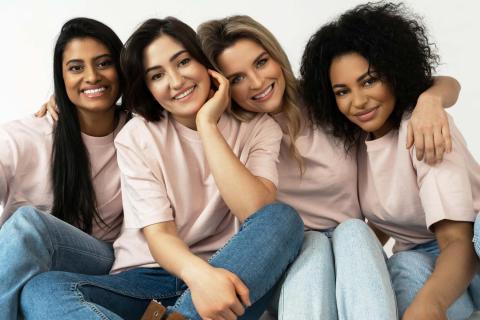 A imagem mostra quatro mulheres sentadas juntas, vestindo camisetas rosa claro e calças jeans. Elas estão sorrindo e abraçadas, transmitindo um forte senso de amizade e união. Cada uma possui traços e estilos distintos, representando diversidade. O fundo é branco, destacando o grupo. A imagem evoca sentimentos de apoio, companheirismo e felicidade.