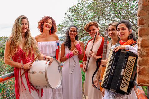 A imagem mostra seis mulheres sorridentes em uma sacada, com árvores ao fundo e uma estrutura de tijolos na lateral. Elas vestem roupas predominantemente brancas com detalhes em vermelho e seguram instrumentos musicais (tambor, triângulo, flauta, violão e sanfona). A cena transmite uma atmosfera leve e artística, sugerindo que fazem parte de um grupo musical. O sorriso e a postura das mulheres indicam alegria e cumplicidade.