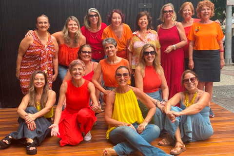 A imagem mostra um grupo de 15 mulheres reunidas em um espaço externo com um piso de madeira. Elas estão vestindo roupas vibrantes em tons de laranja, vermelho e amarelo, e muitas usam óculos de sol. Algumas estão sentadas ou ajoelhadas na frente, enquanto outras ficam em pé atrás. Todas sorriem e posam de maneira descontraída para a foto. O fundo inclui uma parede escura e um ambiente urbano com calçada e vegetação ao fundo.