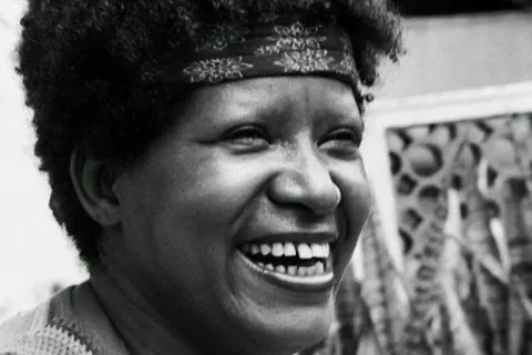 A imagem em preto e branco mostra Lélia Gonzalez sorrindo amplamente. Seu rosto expressa alegria, com olhos levemente semicerrados e dentes à mostra. Seu cabelo crespo é adornado por uma faixa estampada na testa. O fundo apresenta elementos arquitetônicos vazados e algumas plantas, sugerindo um ambiente externo ou uma varanda. A luz suave realça os contornos do rosto, destacando sua expressão vibrante e acolhedora.