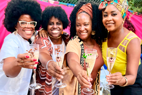 A imagem mostra quatro mulheres negras sorridentes, segurando taças com espumante em um brinde. Elas estão vestidas com roupas coloridas e estampadas, algumas usando turbantes e acessórios vibrantes. O fundo tem um tecido rosa forte, sugerindo uma decoração festiva. A expressão de felicidade e a proximidade entre elas indicam um momento de celebração e confraternização.