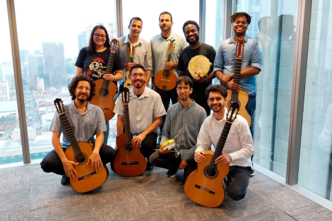 A imagem mostra um grupo de nove músicos posando em frente a uma janela com vista para prédios urbanos. A maioria segura violões, enquanto dois seguram pandeiros. Eles vestem roupas casuais e sociais, e exibem expressões sorridentes. A iluminação natural realça o ambiente moderno.