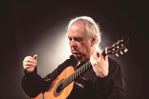 A imagem mostra o violonista Edelton Gloeden em um momento de concentração enquanto toca seu violão. Com expressão serena e gestos precisos, ele veste uma camisa preta e está envolto por uma iluminação suave que destaca sua presença e cria um clima intimista. A cena transmite a sensibilidade e a profunda conexão de Gloeden com a música erudita e o violão.