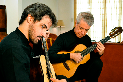 Os músicos que formam o Duo Roberto Victório e Sergio Ribeiro, estão sentados em um ambiente interno, tocando violões com concentração e sensibilidade. Ambos vestem roupas escuras. Um deles tem barba e cabelo escuro e cacheado; o outro tem cabelo grisalho. A cena transmite um momento íntimo de execução musical, com luz natural entrando por uma janela ao fundo e criando uma atmosfera acolhedora.
