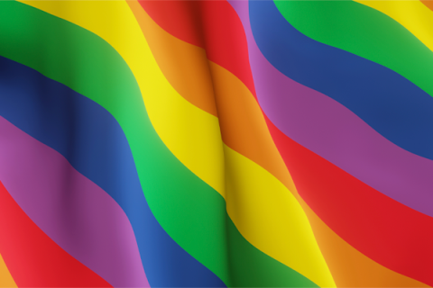 Imagem de uma bandeira LGBTQIA+ estilizada, com faixas onduladas em seis cores vibrantes: vermelho, laranja, amarelo, verde, azul e roxo. As ondas dão movimento ao tecido, criando a sensação de leveza e fluidez. A bandeira simboliza o orgulho, a diversidade e a luta pelos direitos da comunidade LGBTQIA+.