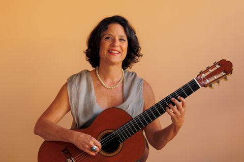 A violonista Adriana Ballesté aparece sorridente, segurando um violão em posição de toque diante de um fundo em tom pêssego. Usa um vestido claro de tecido leve e colar de pérolas, com os cabelos escuros e ondulados na altura dos ombros. A imagem transmite leveza, elegância e afinidade com a música, refletindo uma atmosfera acolhedora e serena.
