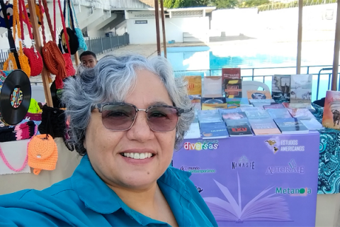Mulher de cabelos grisalhos e curtos, usando óculos escuros e camisa azul, sorri para a câmera em uma selfie. É Léa Carvalho, publisher da Editora Metanoia, especializada em Gênero, Sexualidade e Diversidade. Ao fundo, há uma mesa com diversos livros expostos sobre uma toalha roxa com palavras como “diversas”, “Metanoia” e “Autor(a)Me”. Também há bolsas coloridas penduradas à esquerda e uma piscina ao fundo, sugerindo um evento ao ar livre, como uma feira cultural ou literária.