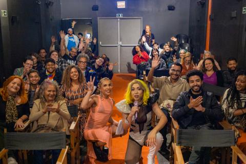 Pessoas de diferentes idades e estilos estão sentadas em um auditório pequeno, sorrindo e acenando para a câmera. Na frente, três pessoas se destacam, incluindo uma drag queen com peruca loira e figurino chamativo, que segura um microfone. O ambiente é acolhedor e descontraído, sugerindo um evento cultural ou artístico com forte senso de comunidade e celebração da diversidade.