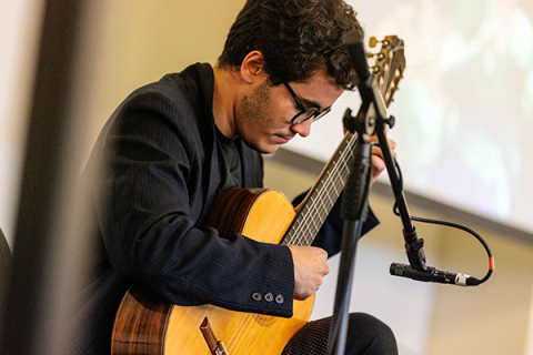 O violonista Tomas Haushahn (RS/EUA) toca com concentração, sentado diante de um microfone. Usa óculos, blazer escuro e camiseta preta. A imagem capta o gesto preciso de sua performance musical, com destaque para o violão e a expressão focada do artista. Ao fundo, uma tela desfocada sugere que ele se apresenta em um evento cultural ou recital.
