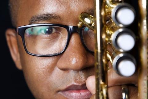 A imagem mostra um close do rosto do clarinetista Everton Machado, que usa óculos e tem expressão séria e concentrada. Ele segura um clarinete de metal dourado próximo ao rosto, com os pistões destacados em primeiro plano. A iluminação foca seu olhar e o instrumento, criando uma atmosfera de intensidade e conexão com a música. O fundo escuro realça os detalhes do instrumento e a expressão do músico.